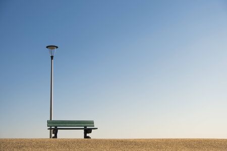 abandoned resting bench with lamppost mounted on the asphalt with copy space against a blue sky, loneliness conceptの写真素材