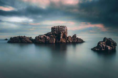 Beautiful long exposure against dramatic sunset sky on the beach with dragon rock and calm waterの写真素材