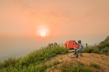 man sitting and drinking coffee near camping tent with sunrise or sunset backgroundの写真素材