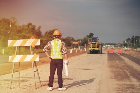 Engineer wear a hard hellmet and holding blueprint on road construction site with machinery,safety first conceptの写真素材