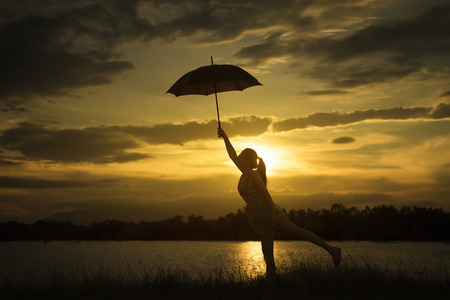 woman holding Umbrella at sunset moment near lake.cloudy sky and big mountain in background.silhouette.jump with umbrella at sunsetの写真素材