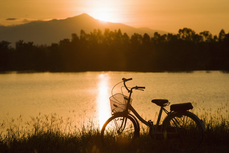 retro vintage bicycle near the lake at sunset moment. silhouette bicycle at the sunset with grass field.big mountain and sunset background.journey concept.lonely conceptの写真素材