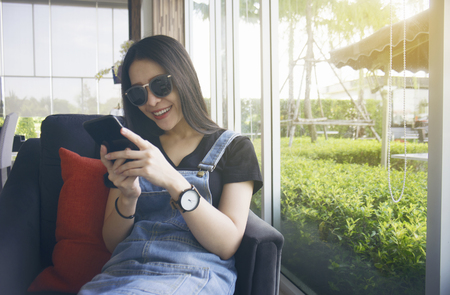 Cute young asian woman using a smart mobile phone while sitting in the lobby of hotel in fresh morning dayの写真素材
