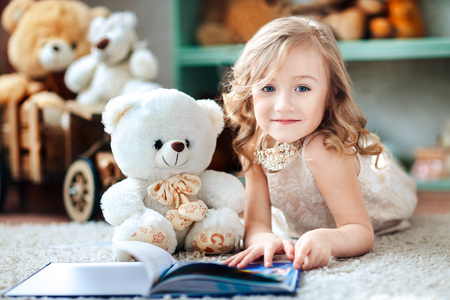 Little blonde girl is reading a book in a children's room with a toy teddy bearの写真素材
