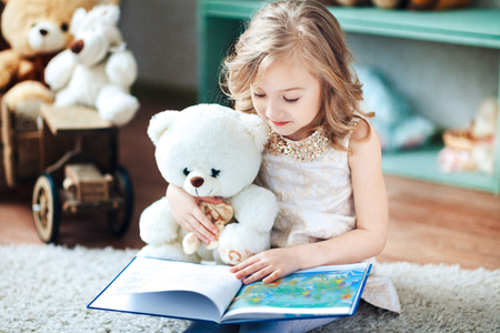 Little blonde girl is reading a book in a children's room with a toy teddy bearの写真素材