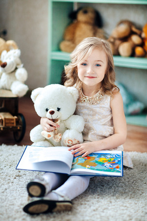 Little blonde girl is reading a book in a children's room with a toy teddy bearの写真素材