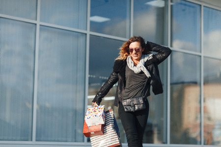 Beautiful girl in sun glasses is holding shopping bags, looking at camera and smiling while standing outdoorsの写真素材