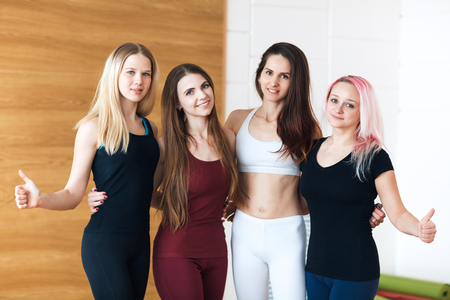Group of young sporty girls showing thumbs up while standing at white interior of loft studio. Female companions in gym resting after fitness, indoorの写真素材