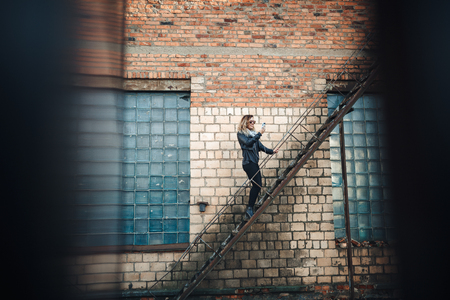 Smiling young woman in sunglasses, a black leather jacket, black jeans standing on an urban metal stair against a brick wall and dances. Woman listening to music on the stairs of industrial building.の写真素材