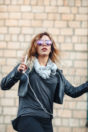 Young beautiful sexy woman with headphones and smartphone in hand in mirrored sunglasses, a black leather jacket, black jeans dancing by the wall of the industrial building of brick on the street.の写真素材