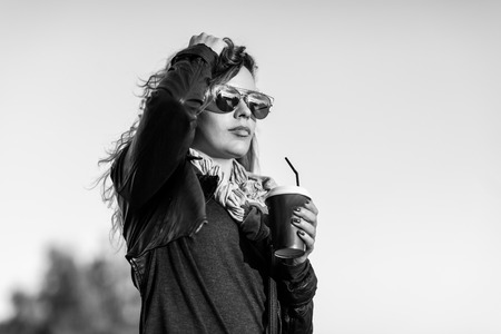 Attractive woman in mirrored sunglasses, a black leather jacket, drinking coffee on the waterfront river in the city in front the setting sun. Waving hair. city,bridge. Black and white pictureの写真素材
