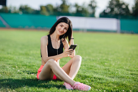 Young sporty joyful woman brunette in shorts and top sitting on the grass football field stadium and listens to music in earphones, her head thrown back, she gets pleasure, relaxes.の写真素材