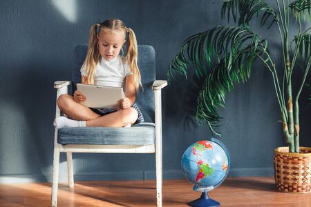 Pretty smiling blondy girl relaxing on a chair near the globe indoors at home with a tablet pc in her socks and jeansの写真素材