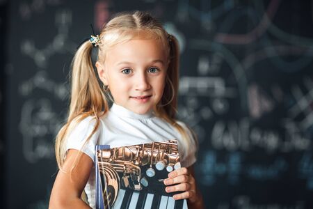 Smiling little blonde girl with hair gathered in tails, white t-shirt, and gray skirt is holding...
