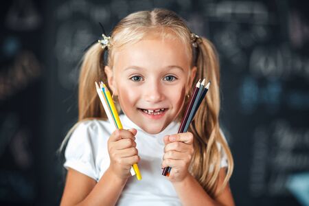 Back to school! Portrait a little smiles girl stands with girl holding colored pencils near the face against chalkboard with school formulas at schoolの写真素材