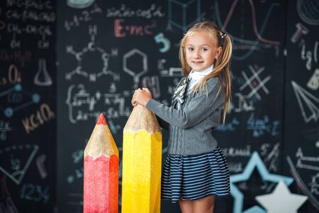 Back to school! A little blonde girl in school uniform stands with two very large red and yellow pencils on the floore against chalkboard with school formulas at school. The kid is studying in the classroom.の写真素材