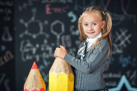 Back to school! A little blonde girl in school uniform stands with two very large pencils on the floore against chalkboard with school formulas at school. The kid is studying in the classroomの写真素材