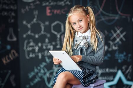 Adorable little blond schoolgirl in school uniform holding white digital tablet. Chalkboard with school formulas background. Back to school conceptの写真素材
