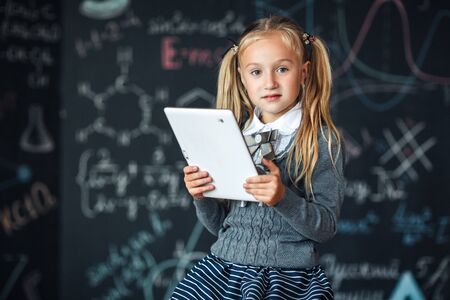 Little blond Girl in school uniform holding white tablet pc in chemistry class. Chalkboard with school formulas backgroundの写真素材
