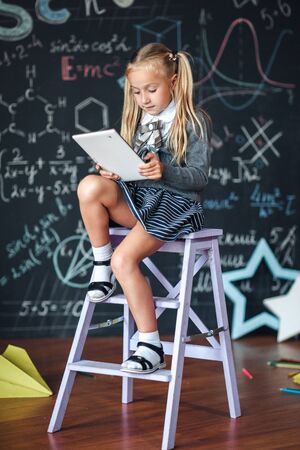 Little blond Girl in school uniform holding white tablet pc in chemistry class. Chalkboard with school formulas backgroundの写真素材