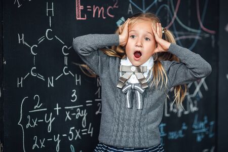 Little girl blonde in school uniform holding hands on her head., open his mouth against chalkboard with school formulas. Complex school program, does not match the age of the childの写真素材