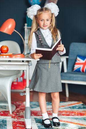 schoolgirl blondy in school uniform with white bows standing near the table and studying at home. Education and school conceptの写真素材