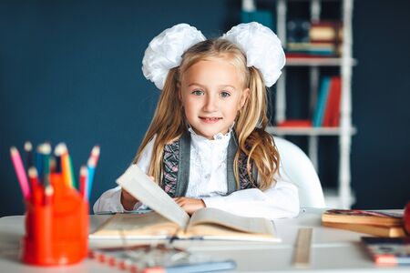 Child and school supplies home Schoolgirl at the table with a textbook and colorful stationery. Girl in school uniform at a Desk in the classroom, holding a textbook, looking at the camera and smiling.の写真素材