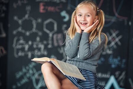A sweet blond girl schoolgirl sits on a chalkboard with school formulas background. Works homework...