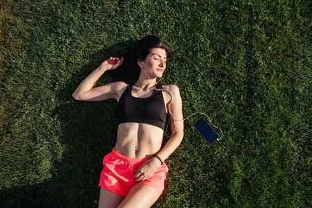 Portrait of a happy young fitness brunette woman laughing and resting on grass and listening to...