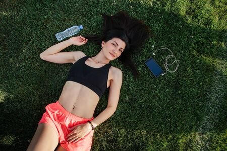 Portrait of a happy young fitness brunette woman lying on the football pitch with a bottle of water...