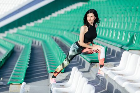 Portrait of a young sports woman leggings and black top near the green and white seats stretcing legs on the stairs before morning jogging in the modern stadium. Healthy lifestyle in the city.の写真素材