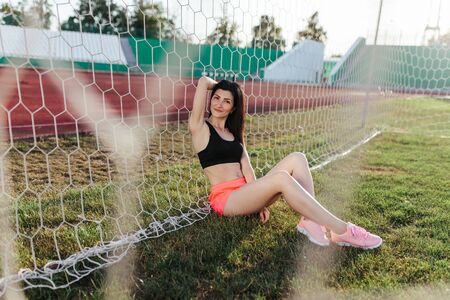 Beautiful stylish brunette woman in black top and pink shorts sitting on the lawn near a football goal at the stadium at sunset. Beautiful sunlight. A girl with a perfect figure and in great shape.の写真素材