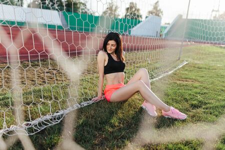 Beautiful stylish brunette woman in black top and pink shorts sitting on the lawn near a football goal at the stadium at sunset. Beautiful sunlight. A girl with a perfect figure and in great shape.の写真素材