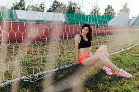 Beautiful stylish brunette woman in black top and pink shorts sitting on the lawn near a football goal at the stadium at sunset. Beautiful sunlight. A girl with a perfect figure and in great shape.の写真素材