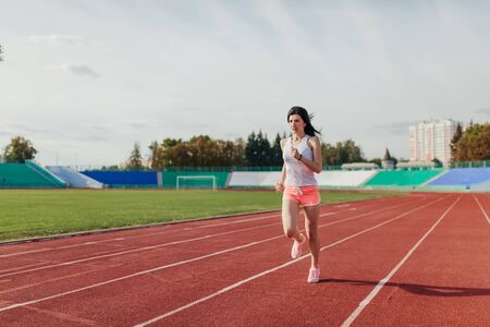 Full body of girl running track on stadium. Real front view of young woman in pink shorts and tank top and pink sneakers. Outdoors, sport.の写真素材