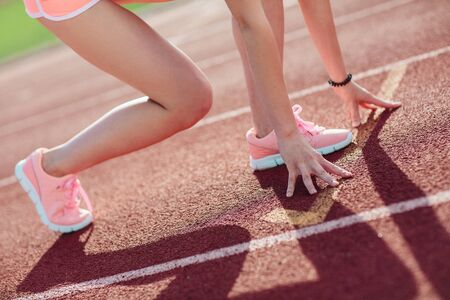 side view Athletic woman in pink shorts and tank tops on running track getting ready to start run, Amateur athlete. Close-up legs and arms.の写真素材