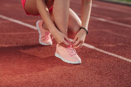 Young woman hands tie laces on her pink sport shoes on a stadium on a running path. White stripe near her. Close-up of tying shoes.の写真素材
