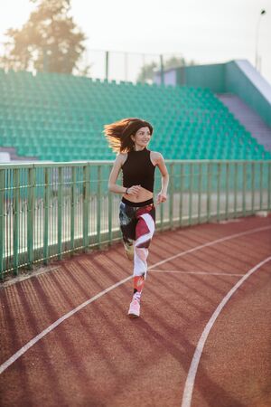 Sport. Athletic young brunette woman in pink sneakers, leggings and top run on running track stadium at sunset. her hair is developing. Concept run. concept of a healthy lifestyleの写真素材