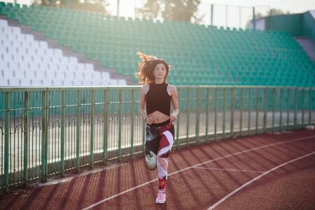 Sport. Athletic young brunette woman in pink sneakers, leggings and top run on running track stadium at sunset. her hair is developing. Concept run. concept of a healthy lifestyleの写真素材