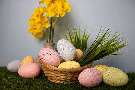 Easter colorful eggs in green grass in a twigs basket with yellow daffodil flowers in the...