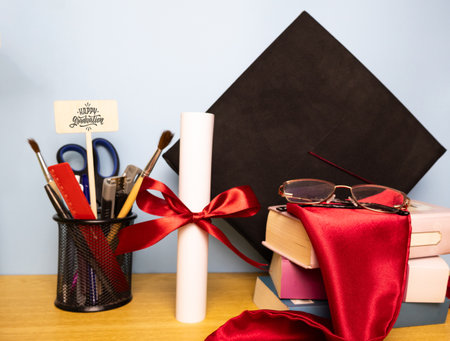 Graduation background with academic hat, pen holder, diploma with red ribbon, eyeglasses, and stole on a stack of booksの写真素材