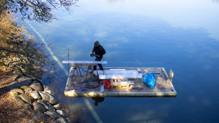 A man working on a platform over a lake, focused on his task amidst the calm water and natural...