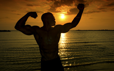 Silhouette of bodybuilder posing at the sunrise on the beach, training in the morning, strong man showing his musclesの写真素材