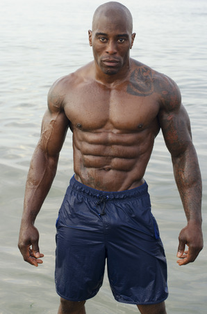 Black bodybuilder relaxing at the beach after a hard workout during a summer vacation. Strong man with perfect abs, pecs shoulders,biceps, tricepsの写真素材
