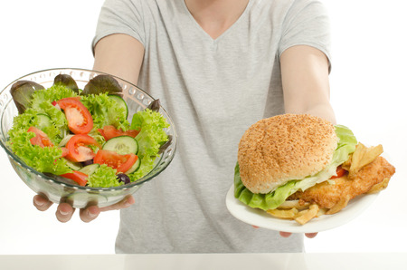 Man offering you a salad and a hamburger. Young man holding in front a bowl of salad and a big burger. Choosing between good healthy food and bad unhealthy food. Fast food versus organicの写真素材