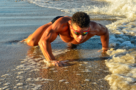 Strong bodybuilder with six pack.Fitness trainer with perfect abs, shoulders,biceps, triceps,chest, flexing his muscles on the beach, training in vacation, doing push ups in the sandの写真素材