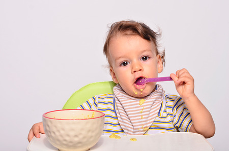 Baby eating food with a spoon, toddler eating messy and getting dirty, infant having oatmeal as breakfastの写真素材
