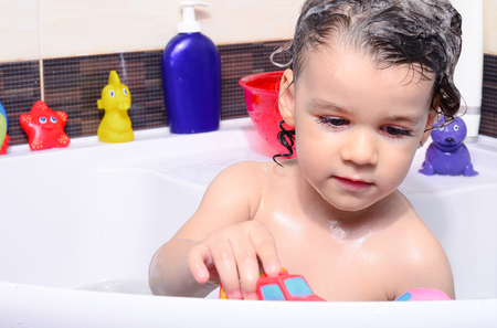 Beautiful toddler taking a bath in a bathtub with bubbles. Cute kid washing his hair with shampoo in the shower and splashing water everywhere. Boy playing with toys in the tubの写真素材