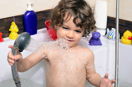 Beautiful toddler taking a bath in a bathtub with bubbles. Cute kid washing his hair with shampoo in the shower and splashing water everywhere. Boy playing with toys in the tubの写真素材