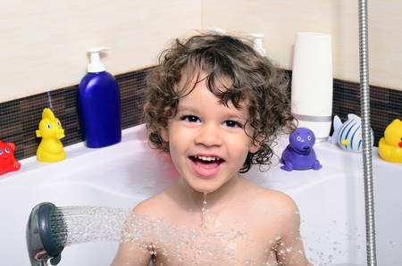 Beautiful toddler taking a bath in a bathtub with bubbles. Cute kid washing his hair with shampoo in the shower and splashing water everywhere. Boy playing with toys in the tubの写真素材